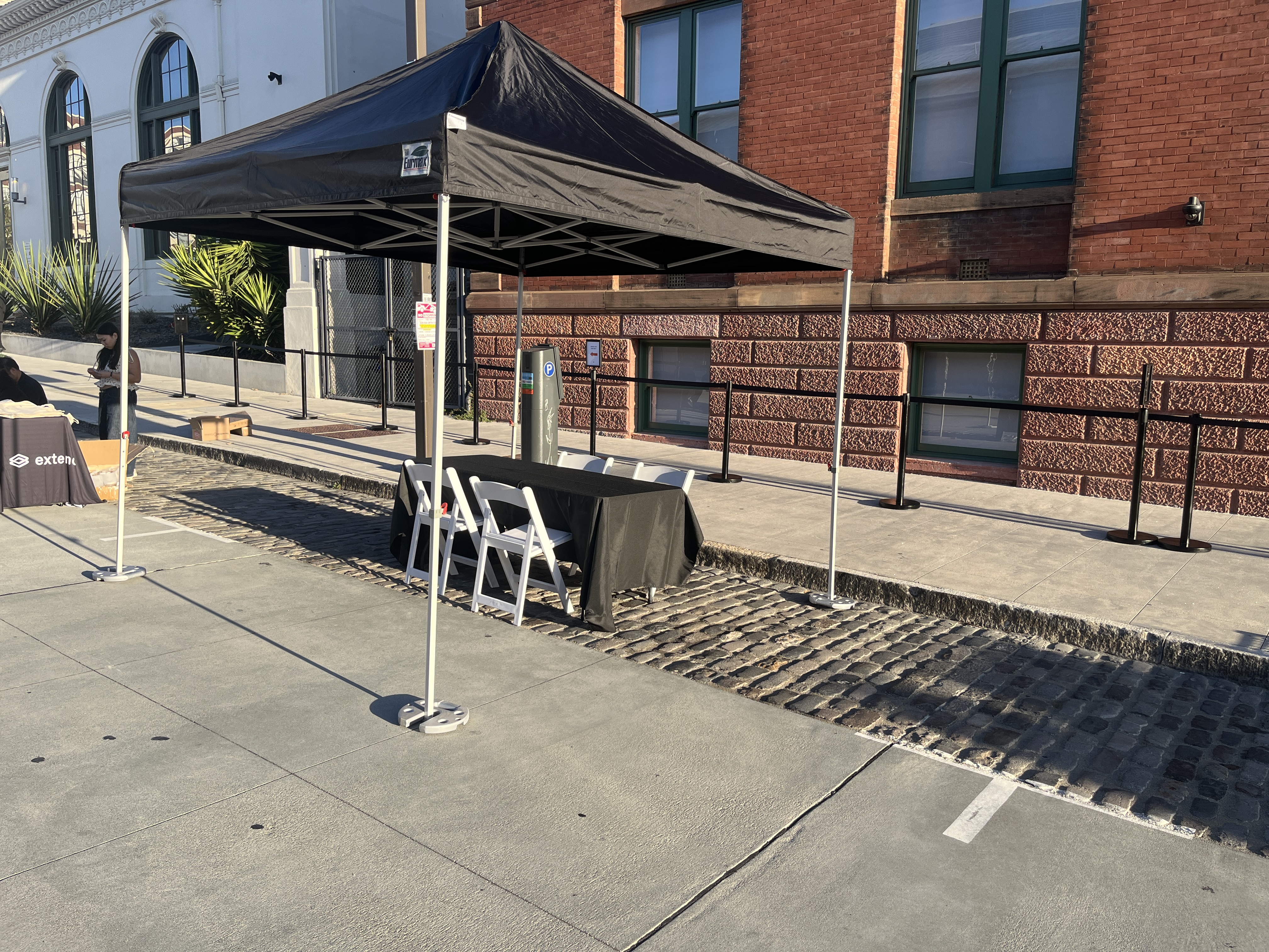 Black pop-up tent, black-clothed table, and white folding chairs set up on a San Francisco sidewalk for Deel's booth at the Y Combinator Alumni Demo Day